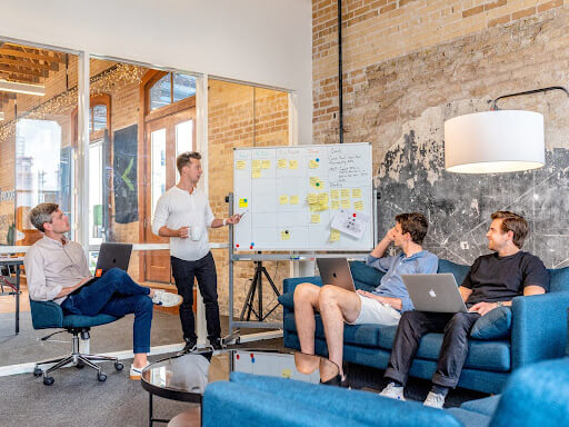 Four social media marketers discuss a content calendar plan in a meeting. One man stands at the whiteboard and points towards it.