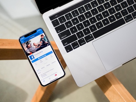 This is an image of a MacBook with an iPhone beside it. The iPhone’s screen displays a Facebook profile.