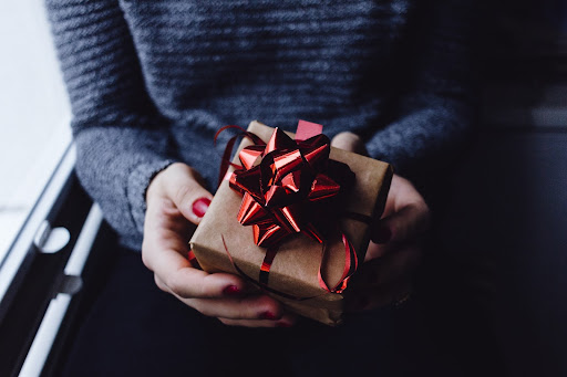 A person with a navy sweater receives a gift covered in brown wrapping and adorned with a red bow. 