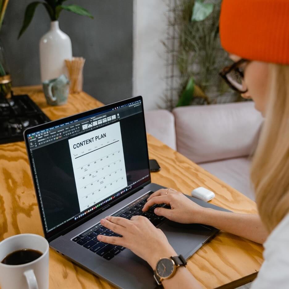 A woman in an orange hat plans a content calendar on her Macbook Pro while she drinks coffee.
