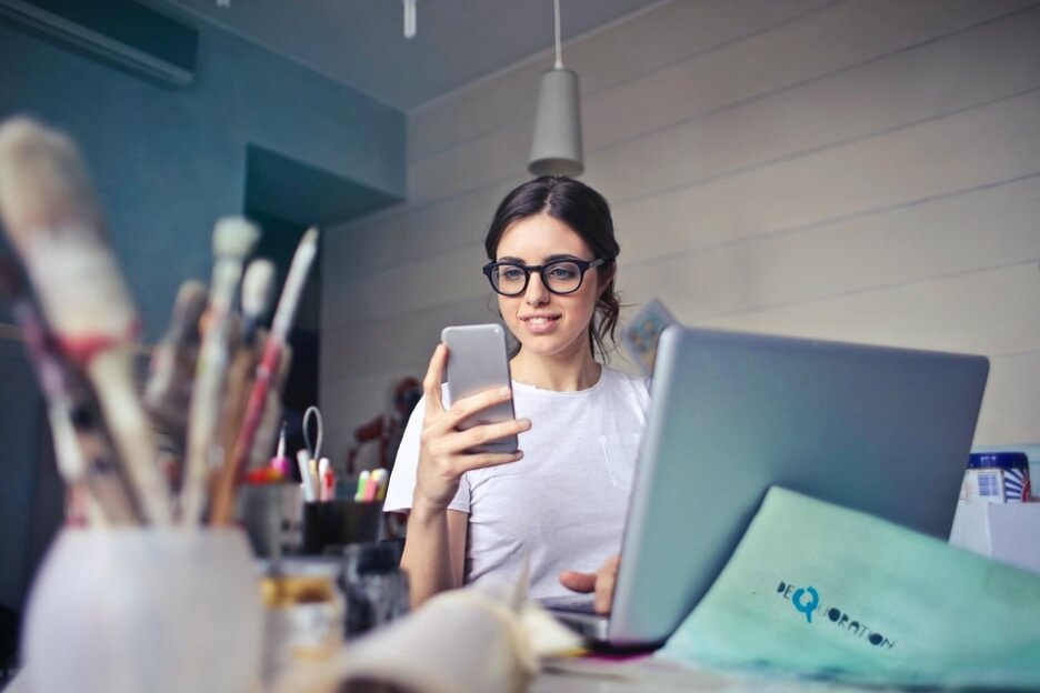 An artist holds her phone in her right hand while she types on her laptop with her left hand.