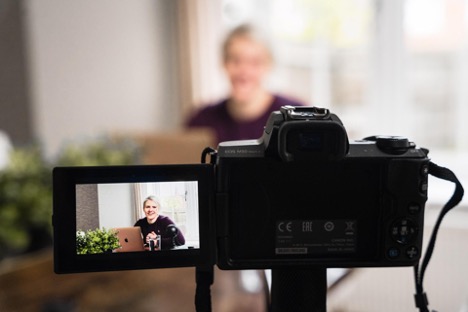 A black Nikon camera films a blonde woman who is seated in front of her MacBook. She addresses the camera.