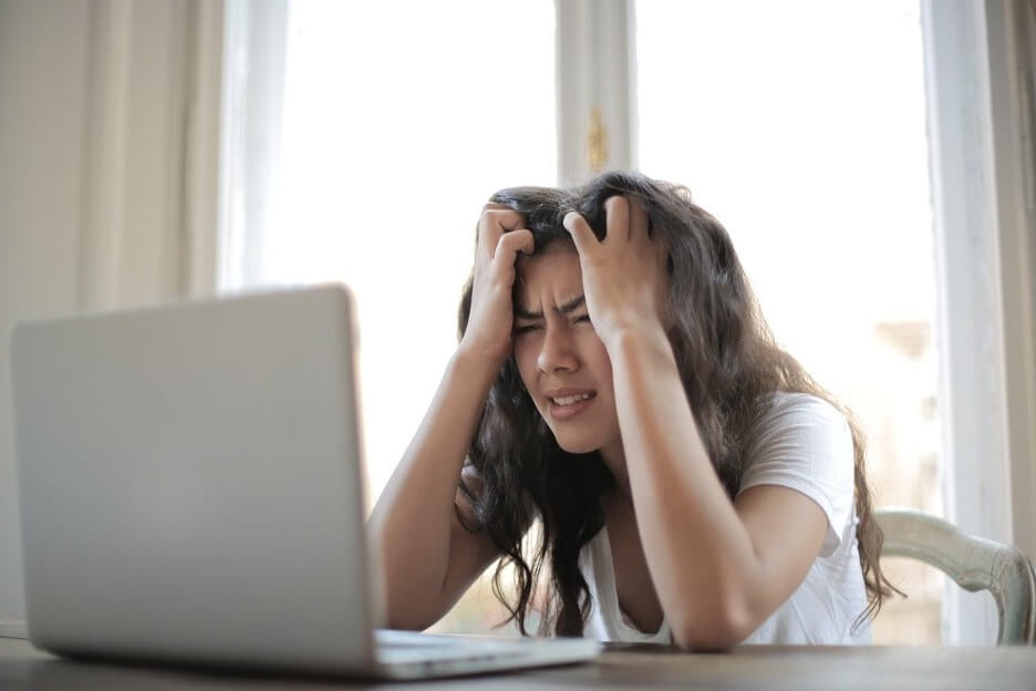 This is an image of a business owner in a white t-shirt who grabs her head as she looks at her laptop.
