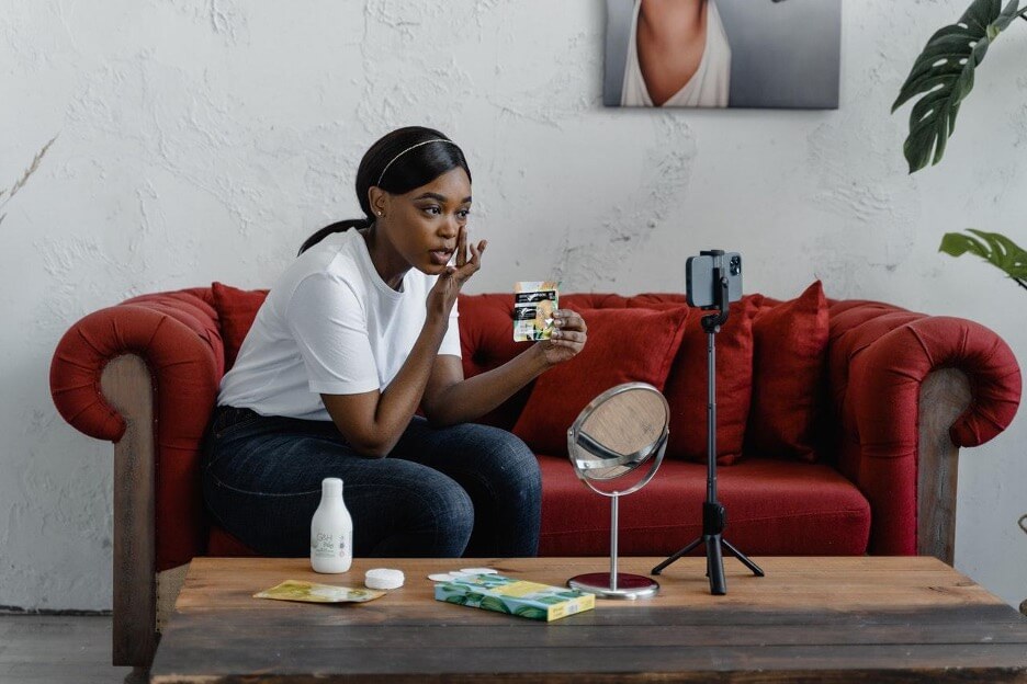 An influencer sits on a red couch while she records a video on how to do makeup.