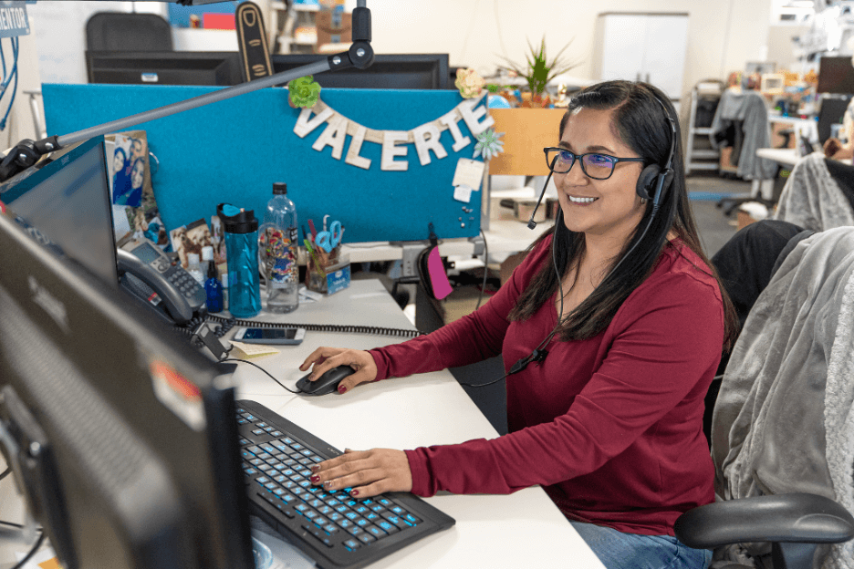 A customer service representative at Zappos smiles as she helps a client over the phone. She wears a headset and glances at the computer screen.