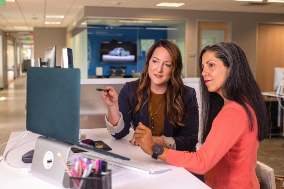 In this image, a digital marketing employee in a navy sweater explains a new strategy to a client wearing a coral shirt.