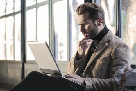 A man wearing a brown jacket holds his chin pensively while he looks at a gray laptop.