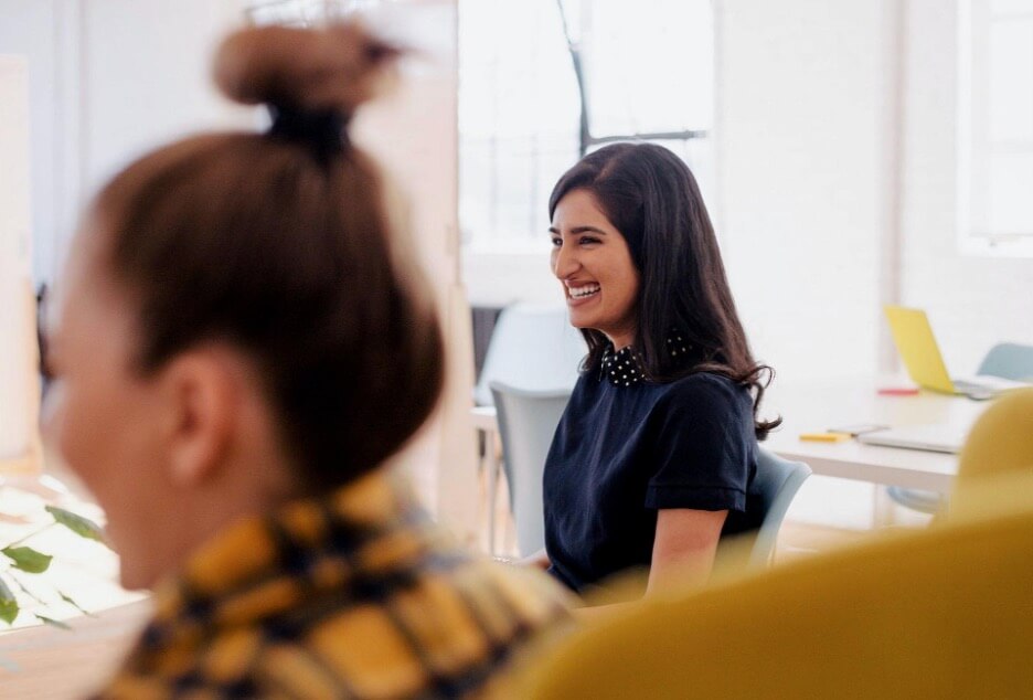 This image shows two female employees in a meeting together. One woman in a navy blue shirt smiles as she sits next to her co-worker in a plaid jacket.