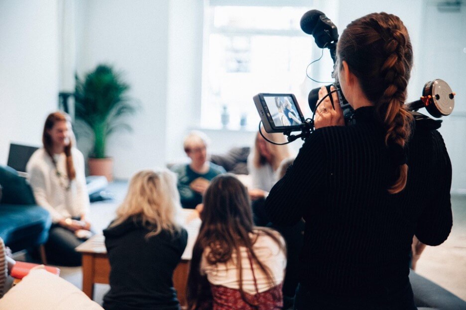 This is an image of a young woman in a black sweater who films a group of people meeting in a living room.