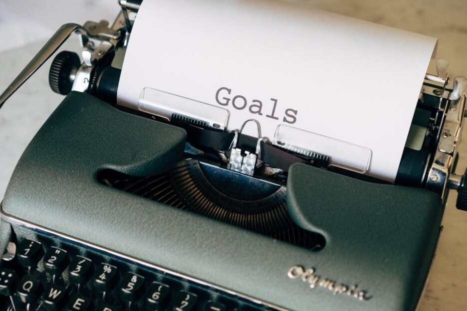 A dark green typewriter sits on a desk and a paper reads “Goals.”
