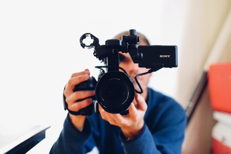 A person with a blue sweater holds a Sony camera and points straight at the camera.