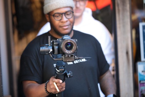 A man in a black t-shirt points a Sony DSLR camera on a gimbal at the person holding the camera.