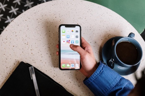 A person wearing a blue shirt uses their iPhone while drinking their coffee.