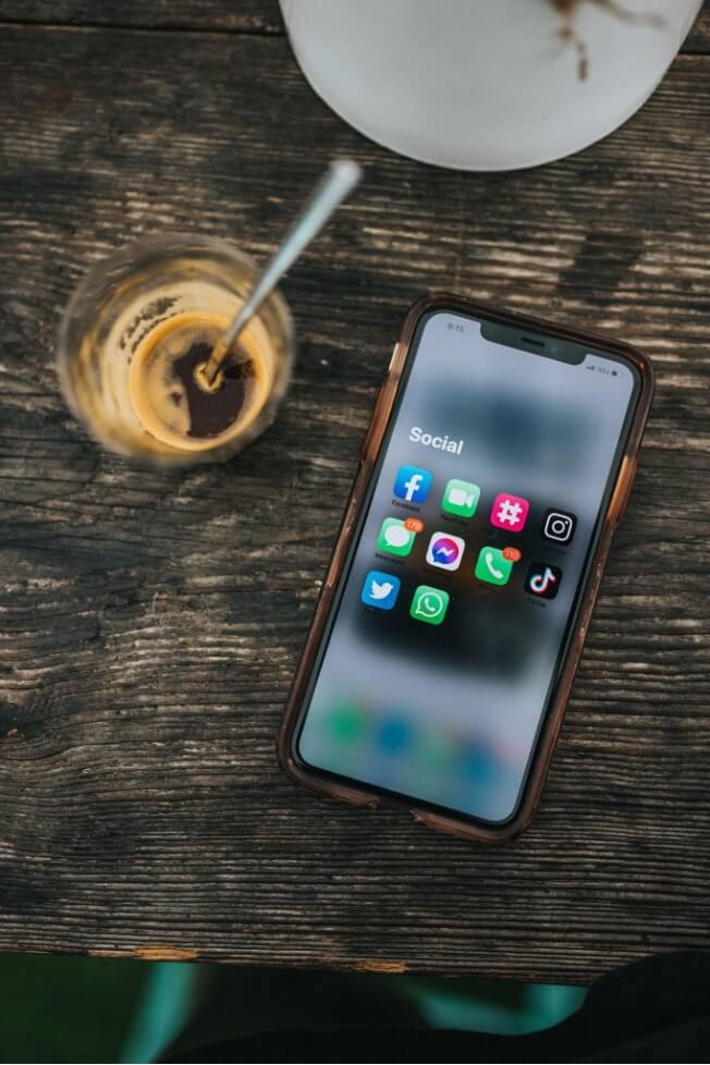 An iPhone resting on a wooden table displays social media apps. A glass of cold coffee is situated beside it.