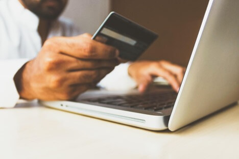 A man in a white shirt looks at his credit card while he types in its number on his MacBook.