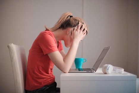 A business owner in a coral t-shirt grabs her head out of frustration while she looks at her laptop.