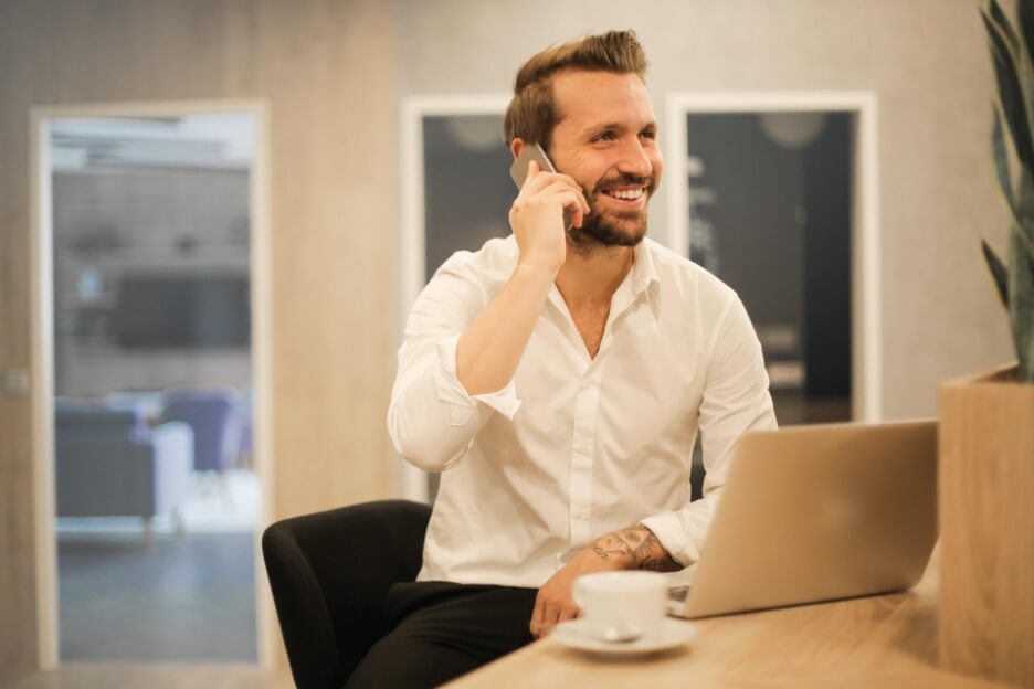 A man in a white button-down shirt talks on the phone while he sits in front of his laptop.