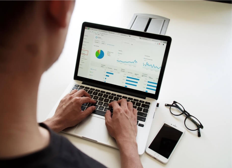 A marketing manager checks the analytics of a client’s social media account and sees how it performs. He wears a black shirt and sits at a white table with his hands typing on a Macbook. An iPhone and glasses are situated next to the laptop.