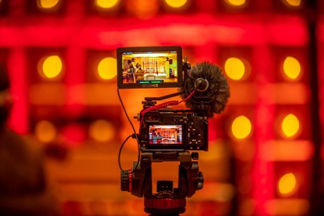 A mirrorless camera on top of a tripod films a red room full of people.