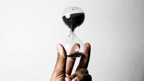 A person holds an hourglass with black sand in their left hand. This is meant to signify the passing of time.