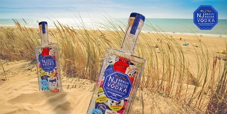 There are two bottles in the sand at a beach. In the background is the ocean. The bottles read: “NJ Beach Badge Vodka.”