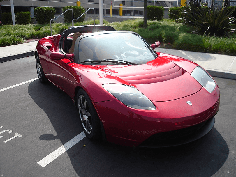This is an image of a red Tesla Roadster from 2006. It sits in a parking lot.