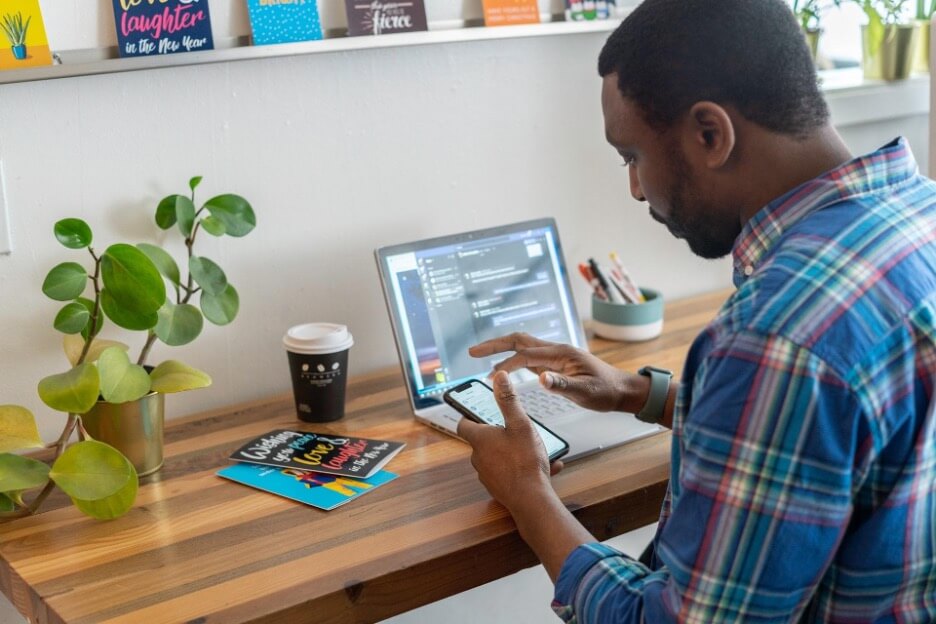 A man in a blue plaid shirt looks at his iPhone while a laptop sits on his desk.