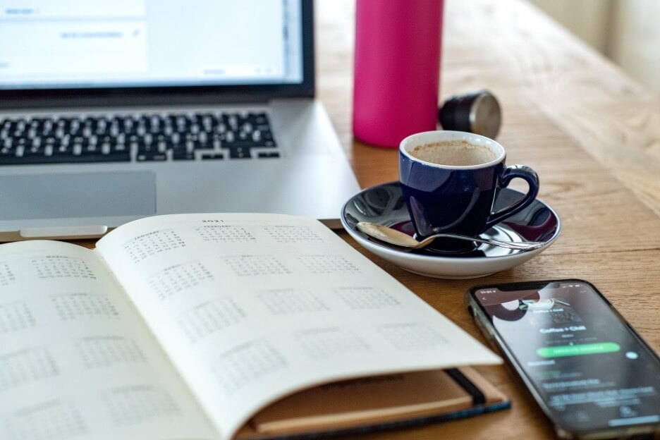 This is an image of a planner on top of a desk, surrounded by a mug, a Macbook, and an iPhone. The planner is open to a page that shows a series of calendars.