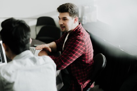 A social media marketing employee in a red plaid shirt explains a strategy to his coworker dressed in a white shirt.