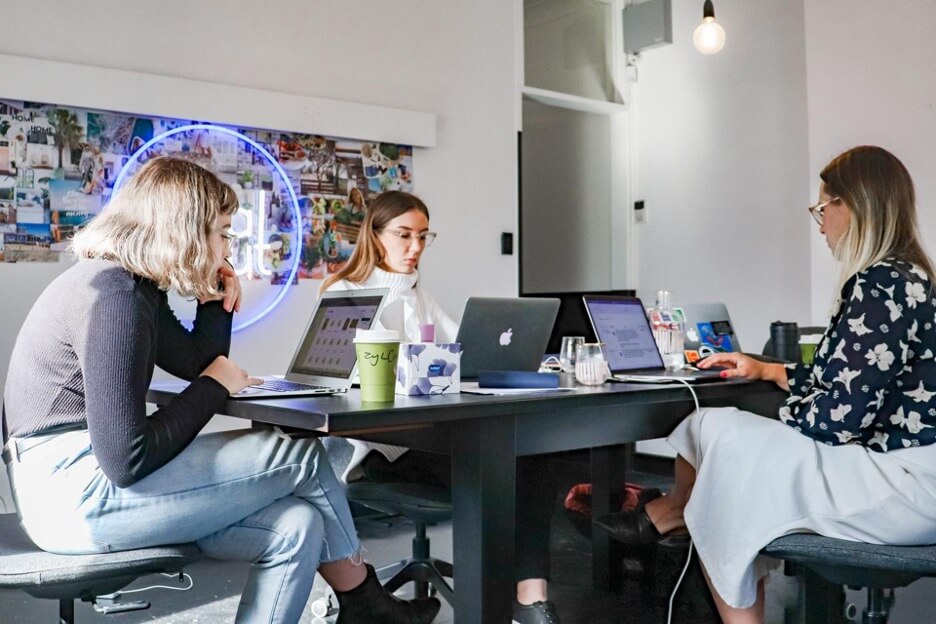 Three social media marketers meeting to work on tasks for a new client. All three look down at their laptops as they discuss the details.