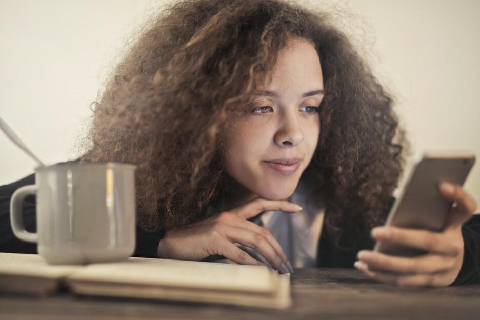 A woman looks at her phone while she eats out of a soup cup.