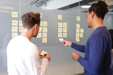 A man in a blue shirt places a sticky note on a glass wall while another man in a white shirt jots down a note.